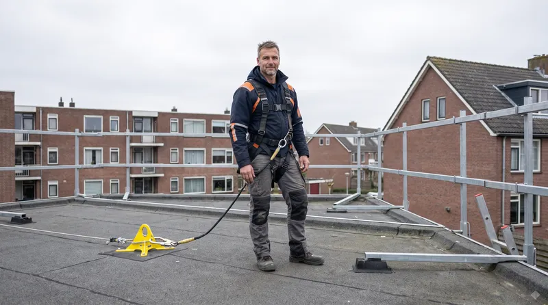 Valbeveiliging met dakanker en randbeveiliging Dakdekker Bart Veldhuis met een veiligheidsharnas vastgemaakt aan een geel dakanker op een plat dak, met tijdelijke randbeveiliging langs de dakrand